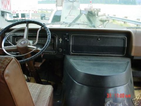 1978 Ford E250 - Interior shot of a full-size van cab looking across the black dash and doghouse engine cover, with a two-spoke steering wheel wearing a lac…