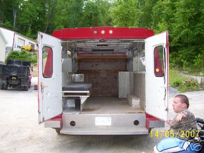 1978 Ford E250 - The rear doors of a red and white van are open, revealing its utilitarian interior with a bench seat and metal shelving.