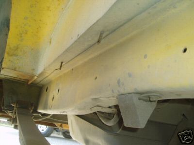 1978 Ford E250 - Underside close-up along a van's rocker panel and frame rail, showing pale yellow-tinted undercoating over the sheet metal with drilled hol…