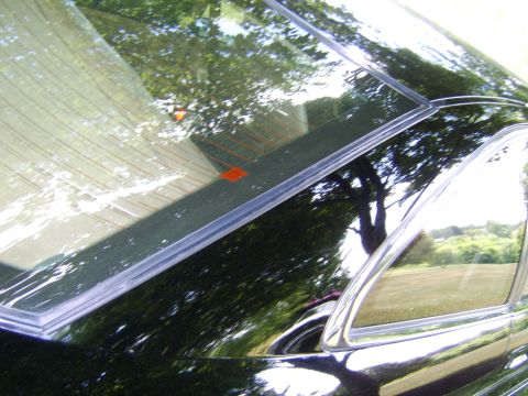 1996 Mercury Grand Marquis - Close-up of a black vehicle's rear window and C-pillar area, showing the glass with defroster lines and an orange decal, plus chrome trim m…