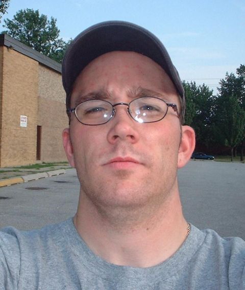 1985 Ford Bronco - A man wearing glasses and a baseball cap poses for a selfie in an outdoor setting with a car visible in the distance.