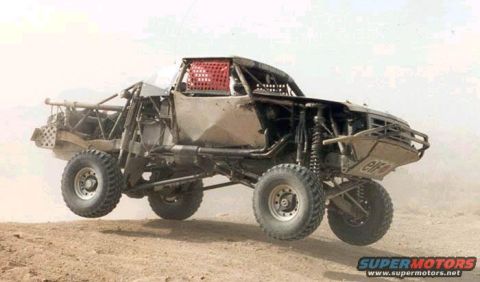 1990 Ford Bronco - stripped tube-frame off-road race truck catches air over a dusty desert course with a red roll cage net visible.