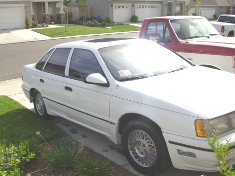 1995 Ford Taurus - A white 1995 Ford Taurus sedan is parked in a driveway, with a red and white pickup truck behind it.