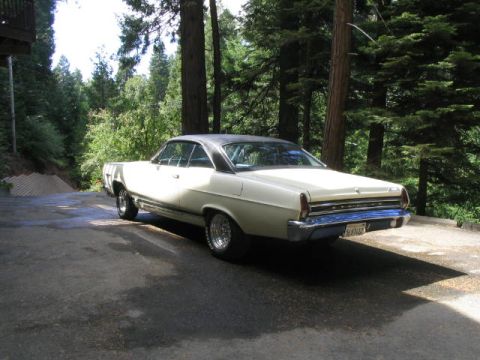 1973 Ford Ranchero - A white coupe with a black vinyl top is parked on a paved surface surrounded by trees.
