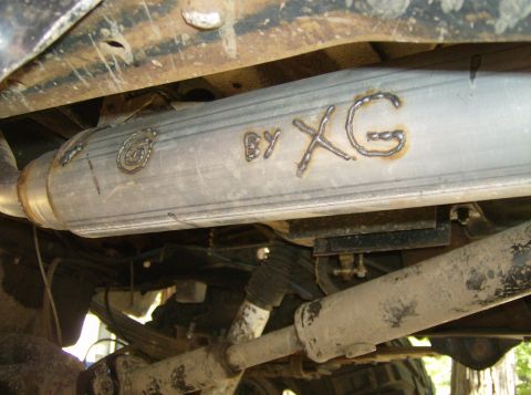 1986 Ford Bronco - Custom exhaust pipe with "BY XG" welded onto it, viewed from underneath a vehicle.