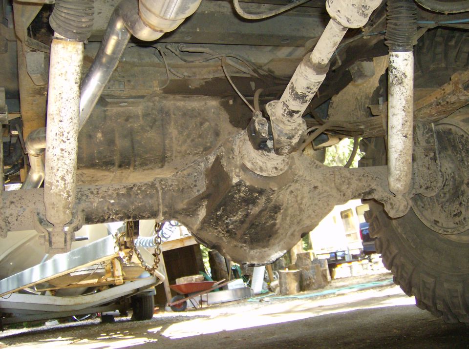Underside view of a vehicle's rear axle, driveshaft, and suspension components. A boat trailer is visible to the left