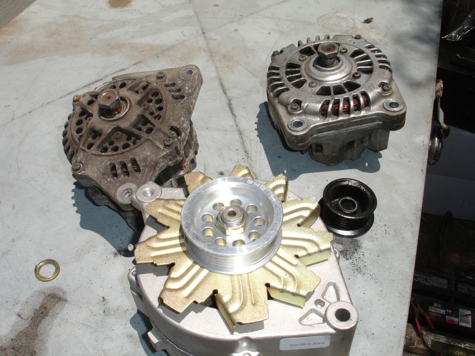 Three alternators and a pulley on a table. One alternator has a gold-colored fan. A black pulley is to the right