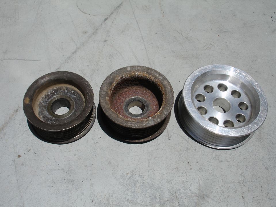 Three automotive pulleys on a gray surface; two are worn and rusty, one is new aluminum