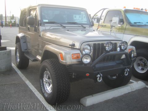 1987 Suzuki Samurai - A tan Jeep Wrangler is parked next to another vehicle, showing its front end with custom modifications.