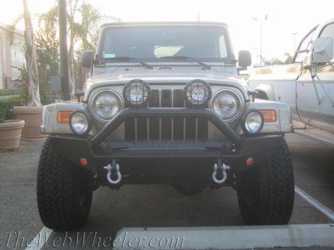 1987 Suzuki Samurai - Front view of a Jeep Wrangler with aftermarket bumper and lights parked next to another vehicle.