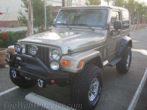 1987 Suzuki Samurai - A tan Jeep Wrangler is parked in a lot with palm trees and a fence in the background.