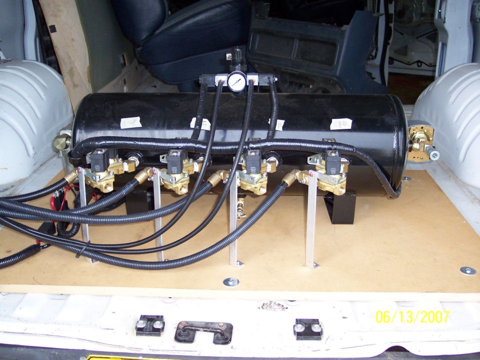 Interior of a van showing a black air suspension tank with multiple solenoid valves and hoses mounted on a wooden platform