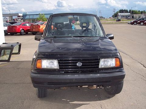 1995 Ford Bronco - A black Suzuki Samurai is parked in a lot, with other vehicles visible in the background.