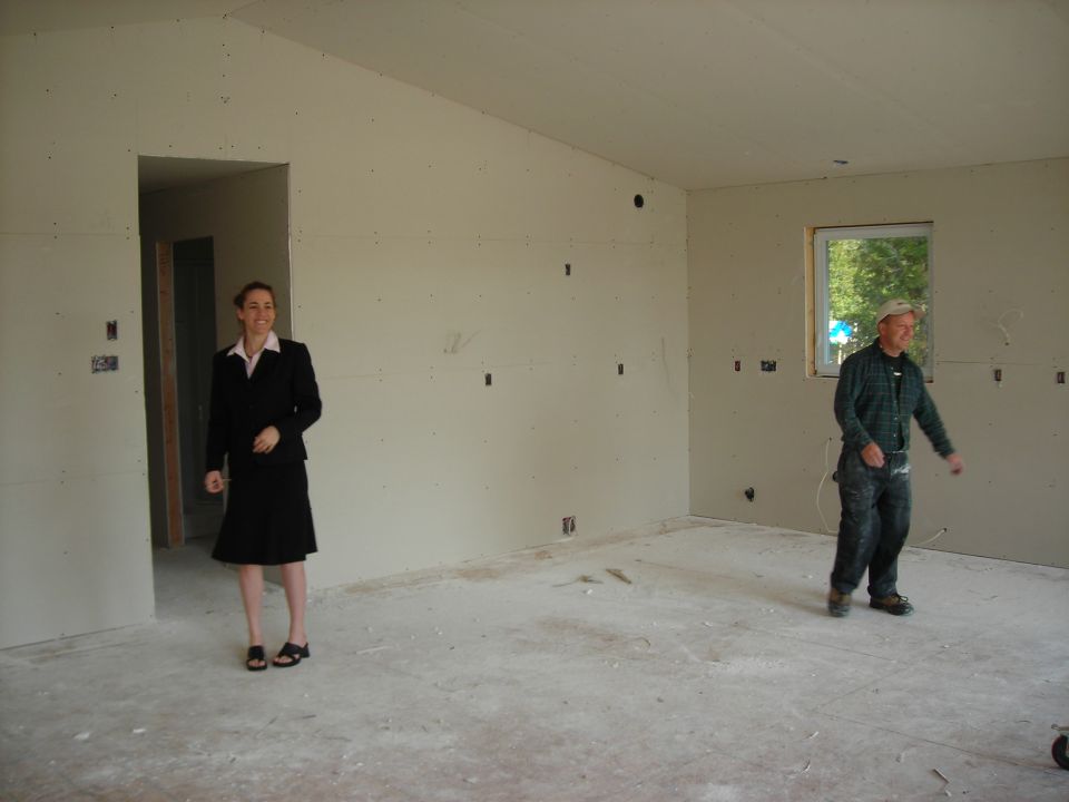 Woman in suit and man in work clothes stand in unfinished room with drywall