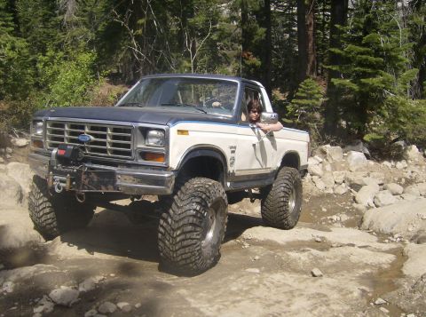 1986 Ford Bronco - A modified Ford Bronco navigates a challenging off-road path surrounded by trees and rocks.