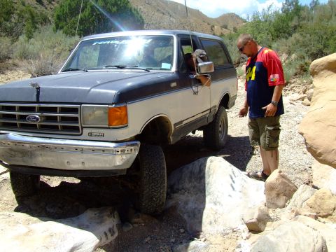 1987 Ford Bronco - A two-tone 1987 Ford Bronco is shown off-roading over large rocks on a sunny day.