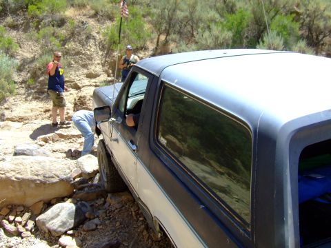 1987 Ford Bronco - A group of people are gathered around a 1987 Ford Bronco as it attempts to drive over a rocky obstacle.
