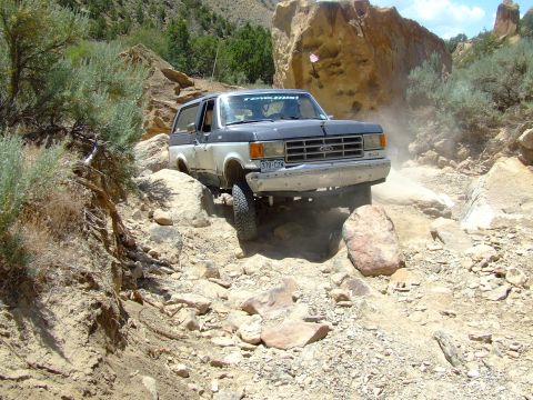 1987 Ford Bronco - A 1987 Ford Bronco tackles a challenging off-road path with large rocks and sparse vegetation.