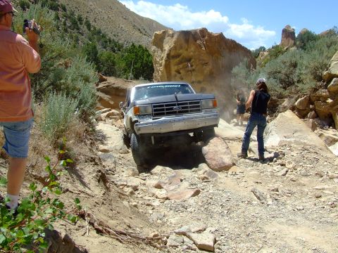 1987 Ford Bronco - A 1987 Ford Bronco tackles a challenging off-road course, kicking up dust as it maneuvers over large rocks.