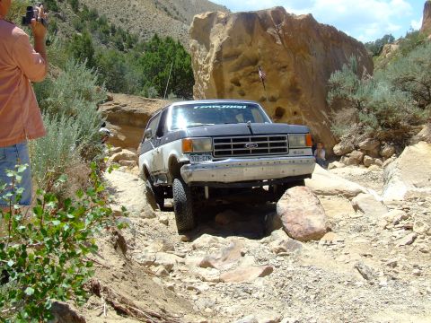 1987 Ford Bronco - A two-tone Ford Bronco is shown driving over a rough, rocky terrain with large boulders and sparse vegetation.