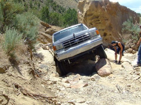 1987 Ford Bronco - A silver 1987 Ford Bronco is shown off-roading on a challenging rocky terrain, with one wheel elevated on a large rock.