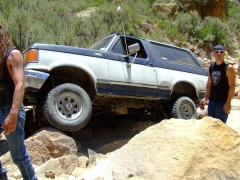 1987 Ford Bronco - A 1987 Ford Bronco is shown off-roading over large rocks, with two men standing nearby.