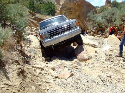 1987 Ford Bronco - A Ford truck is shown attempting to traverse a difficult, rocky off-road path with onlookers present.