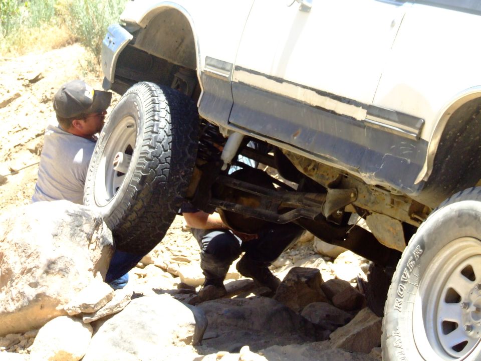 1987 Ford Bronco with front wheel high on rock, people working underneath