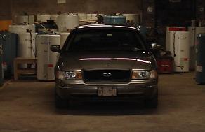 1999 Ford Crown Victoria - Dim head-on front view of a champagne/tan Ford Crown Victoria sedan parked inside a cluttered garage or shop, with a roof-mounted light bar…