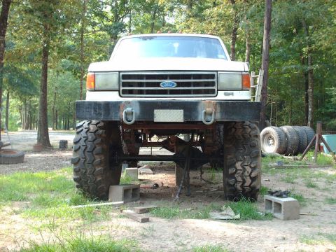 1990 Ford F-150 - Front view of a lifted, white Ford pickup truck with large off-road tires. The truck is parked outdoors in a grassy area with trees in the…