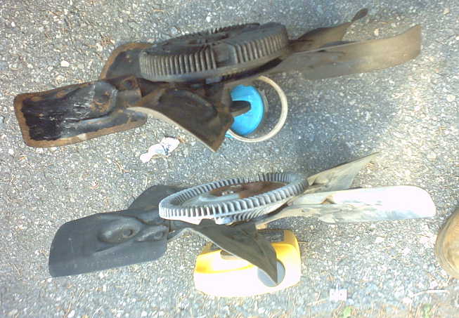 Two engine cooling fans, one black plastic and metal, the other mostly black plastic with a metal hub, resting on gravel