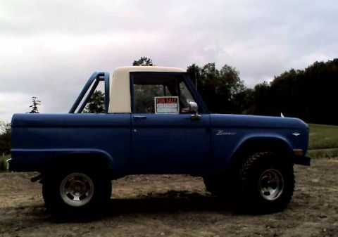 1973 Ford Bronco - A blue 1973 Ford Bronco with a white roof and roll bar is parked on a dirt lot under a cloudy sky.