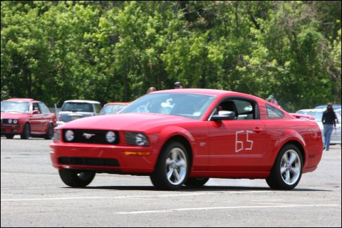 2001 Mercury Grand Marquis - A red Ford Mustang with racing numbers is seen on a track, with other vehicles and trees in the background.