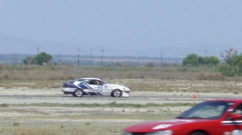 1990 Ford Probe - A race car with blue and white livery speeds around a track, with another vehicle partially visible in the foreground.