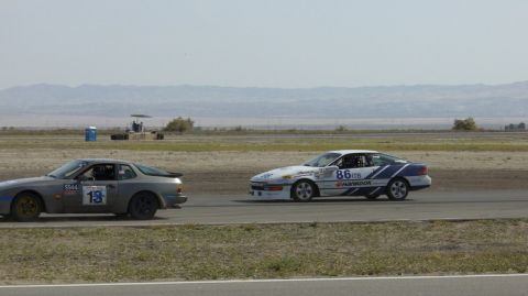 1990 Ford Probe - Two race cars compete on a track under a clear sky with distant mountains.