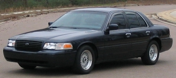 1999 Ford Crown Victoria - Small three-quarter front view of a black Ford Crown Victoria sedan parked on pavement beside a guardrail, shown in a wide letterboxed crop…
