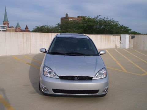 2003 Ford Focus - A silver 2003 Ford Focus hatchback is parked in a lot with a concrete wall and buildings in the background.