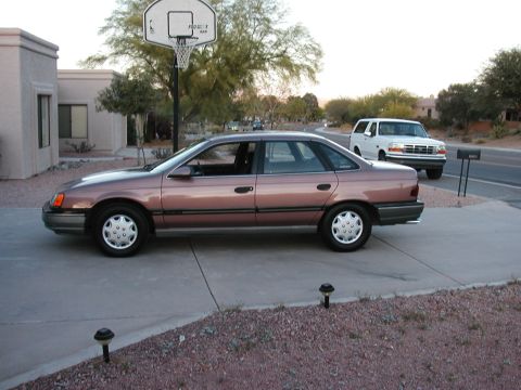 1990 Ford Taurus - Side view of a 1990 Ford Taurus sedan parked on a driveway with a white SUV in the background.