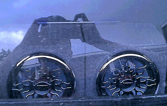 Two chrome flame-pattern subwoofer grilles seen through a vehicle window with reflections
