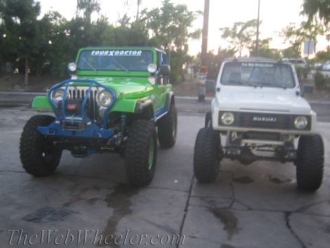 1987 Suzuki Samurai - Two off-road vehicles, a green Jeep and a white Suzuki Samurai, are parked on pavement with trees in the background.