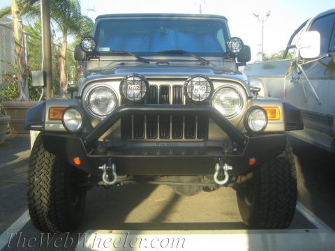 1987 Suzuki Samurai - Front view of a tan Jeep Wrangler, likely from the early 2000s, equipped with KC auxiliary lights and a custom bull bar bumper.