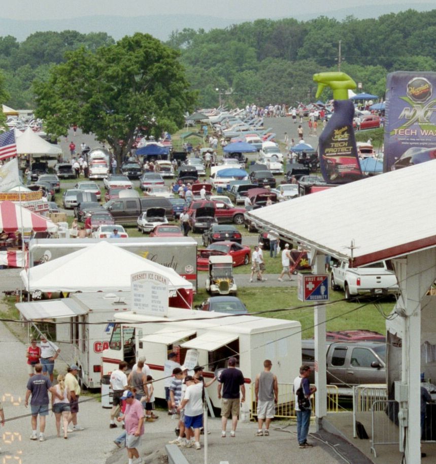 Outdoor car show with many vehicles parked and people milling around vendor tents