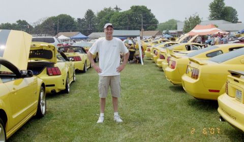 1976 Ford Ranchero - A line of yellow Ford Mustangs are parked on a grassy field, with a man standing in front of them.