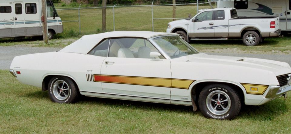 White convertible muscle car with a GT stripe on the side. A white pickup truck and RV are in the background