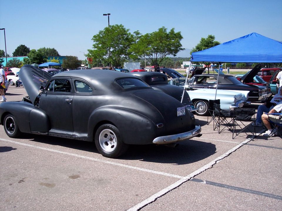 Matte black coupe with hood open parked next to other cars at an outdoor event