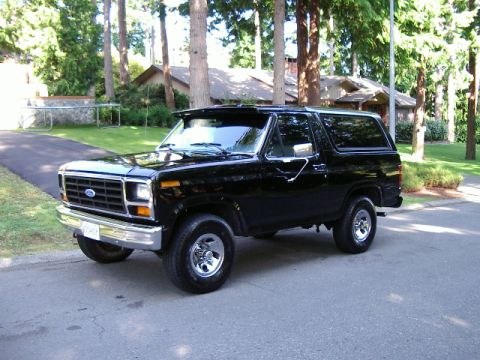 1986 Ford Bronco - A black Ford Bronco is parked on a residential street with trees and houses in the background.