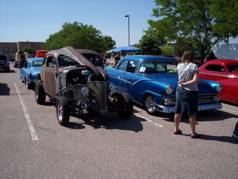 1972 Ford Ranchero - A rusty hot rod is parked with its hood open next to a blue sedan and a red coupe at an outdoor event.