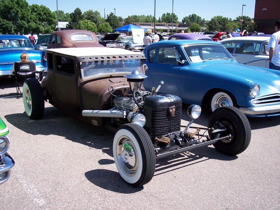 Rat rod with exposed engine and whitewall tires parked next to a blue car