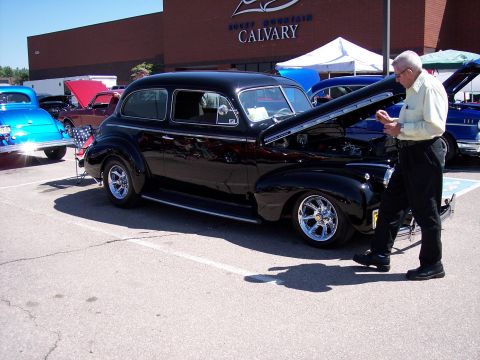 1972 Ford Ranchero - A man inspects a black sedan with its hood up at a car show, with other vehicles visible in the background.