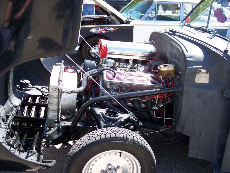 Engine bay of a hot rod with a V8 engine, chrome air cleaner, and headers. Another car with an American flag decal is visible behind it